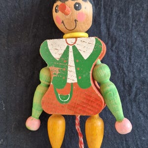Könnte beinhalten: Eine Holzpuppe mit Zugschnur, roter Mütze, grünem Jackett und roter Hose. Die Puppe hat ein bemaltes Gesicht mit einer großen Nase und einem gelben Kragen.