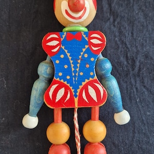 Könnte beinhalten: Eine hölzerne Zieh-Clown-Puppe mit einem rot-blau-weißen Kostüm. Der Clown hat ein bemaltes Gesicht mit einer roten Nase und einen schwarzen Hut mit einem grünen Band.