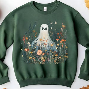 May include: Dark green sweatshirt featuring a whimsical ghost design. The ghost is depicted in a field of colorful wildflowers with butterflies. The design includes orange, yellow, and blue flowers. A fun and unique graphic for Halloween or everyday wear.