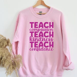 May include: Pink sweatshirt with the text "TEACH compassion TEACH kindness TEACH confidence" in pink.