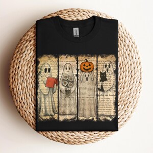 May include: Black t-shirt with a Halloween design. The graphic shows four ghosts, each holding an item: a book, flowers, a pumpkin, and a black cat. The background is made from vintage book pages.