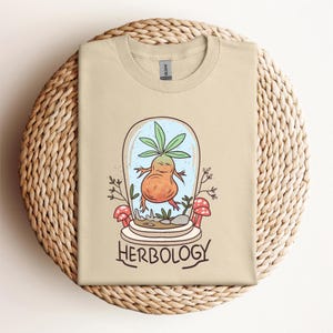 May include: Beige t-shirt featuring a cartoon mandrake plant inside a glass dome with the word "Herbology" below. The mandrake is orange with green leaves and is surrounded by small mushrooms and plants. The shirt is displayed on a woven surface.