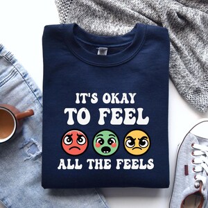 May include: Navy blue sweatshirt with the text "It's okay to feel all the feels" and three cartoon faces expressing different emotions.