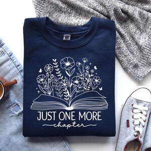 May include: Navy blue sweatshirt with a white design of an open book with flowers and the text "Just One More Chapter".