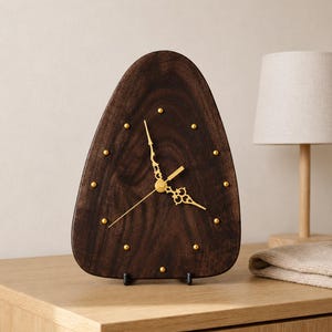 May include: A triangular dark brown wooden clock with a smooth grain finish. The clock face features twelve small gold-coloured metal studs as hour markers and ornate gold-coloured metal clock hands positioned at the centre of the wooden surface.