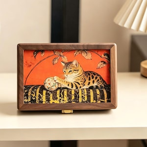 Könnte beinhalten: Holzgerahmte Box mit einer Katzenillustration. Das Kunstwerk zeigt eine braun-beige gefleckte Katze, die mit einem Ball auf einem gestreiften Kissen vor einem orangefarbenen Hintergrund spielt. Der Rahmen ist dunkelbraun.