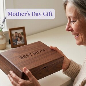 May include: A wooden box with the words "BEST MOM" engraved on the lid. The box is being held by a person. The image also includes a framed photo and the text "Mother's Day Gift".