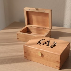 Może przedstawiać: Wyświetlane są dwa drewniane pudełka, jedno otwarte i jedno zamknięte. Zamknięte pudełko ma czarny wzór wieńca laurowego z literą "A" pośrodku. Pudełka wykonane są z jasnego drewna i mają prosty, elegancki design.