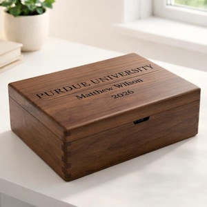 Puede incluir: Una caja rectangular de madera de color marrón oscuro. La tapa está grabada con las palabras "PURDUE UNIVERSITY", "Matthew Wilson" y "2026". La caja está colocada sobre una superficie blanca.