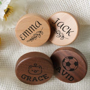 May include: Four wooden round objects with engraved names and designs. Two are light-colored with the names "Emma" and "Jack" and leaf accents. The other two are dark brown, with "Grace" and a bear face, and "David" with a soccer ball.