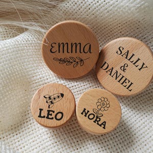 May include: Four small, round wooden objects with engraved designs and text. One has the name "EMMA" with a leaf design. Another says "SALLY & DANIEL". A third has a rocket and the name "LEO". The last one has a flower and the name "NORA".