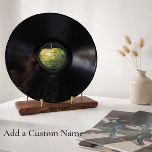 May include: A black vinyl record displayed on a wooden stand with brass supports. The record has a green and yellow label. The stand is engraved with the name "Michael". Additional text reads "Add a Custom Name".