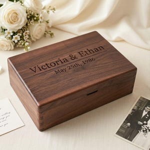 May include: A dark brown wooden box, rectangular in shape, with the names "Victoria & Ethan" and the date "May 25th, 1986" engraved on the lid. The box is set on a cream-colored surface, with a photograph and flowers in the background.