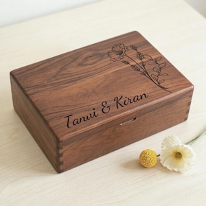 Könnte beinhalten: Eine rechteckige Holzbox mit floralem Design und den Namen "Tanvi & Kiran" auf dem Deckel eingraviert. Die Box hat eine warme braune Farbe und eine natürliche Holzmaserung. Eine kleine weiße Blume und ein gelber Pompon liegen neben der Box.