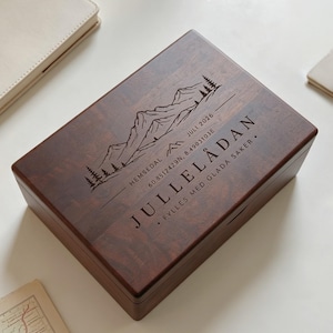 Puede incluir: Una caja rectangular de madera marrón oscuro con tapa abatible. La tapa presenta un grabado láser de una cordillera, pinos y el texto "HEMSEDAL JULI 2026", las coordenadas "60.8512429N, 8.4903103E", "JULLELÅDAN" y "FYLLES MED GLADA SAKER".