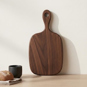May include: A dark brown wooden cutting board with a handle and a hole at the top. The board has a smooth, polished surface and a rounded rectangular shape. A small black cup and a loaf of bread on a linen napkin are in the foreground.