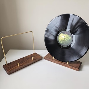 May include: Two wooden record stands with brass accents. One stand holds a black vinyl record with a green and yellow label. The other stand has a brass bar and two brass pegs.