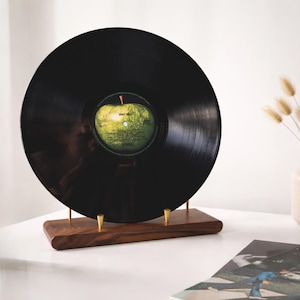May include: A black vinyl record with a green apple label is displayed on a wooden stand with brass supports. The record is a vintage music item, likely an album. The stand is made of dark wood.