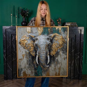 May include: Square framed canvas art featuring a detailed elephant head with gold leaf accents. The elephant is painted in shades of gray, blue, and white, with a textured background of gray and teal. The artwork is held in a gold frame.