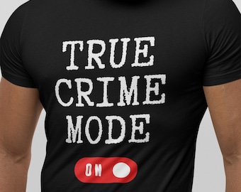 以下が含まれることがあります： 黒いTシャツ。白い文字で「TRUE CRIME MODE」と書かれています。その下に「ON」という文字と白い丸がある赤いグラフィックがあります。