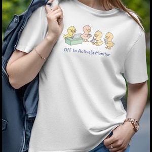 May include: White t-shirt featuring a cartoon graphic of four yellow ducklings working on a laptop, drinking coffee, and taking notes. The text "Off to Actively Monitor" is printed below the graphic. The shirt is paired with blue jeans.