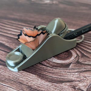 May include: A miniature, gray, 3D-printed pencil plane with a pencil and wood shavings. The pencil plane has a small, round wheel and a blade. The wood shavings are orange and black. The pencil is black.