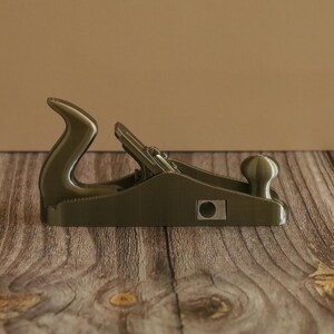 May include: A dark green hand plane, a woodworking tool, is displayed on a wooden surface. The plane has a curved handle and a knob at the front. The tool is designed for smoothing and shaping wood surfaces.