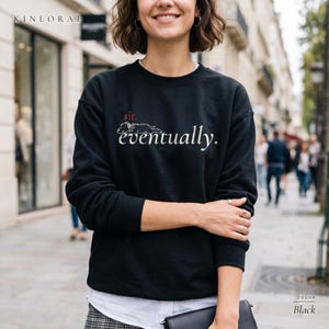 May include: A black long-sleeve crewneck sweatshirt featuring a graphic of a dog lying down with the red text "sit." above the white text "eventually." The sweatshirt has a ribbed neckline, cuffs, and hem, and is worn over a white collared shirt.