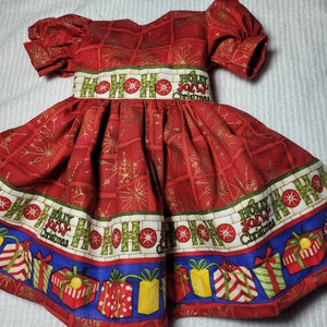 May include: Red Christmas dress with a white and green "HO HO HO" and "Holly Jolly Christmas" print. The dress has a gathered skirt and puff sleeves.