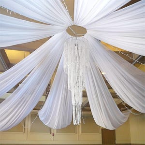 May include: A white fabric ceiling installation with a central chandelier. The fabric is draped in flowing lines from a central point, creating a tent-like effect. The chandelier is a multi-tiered design with hanging crystal beads.