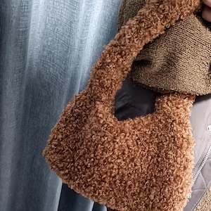 A person holding a brown curly teddy fleece handbag against a blue curtain backdrop.