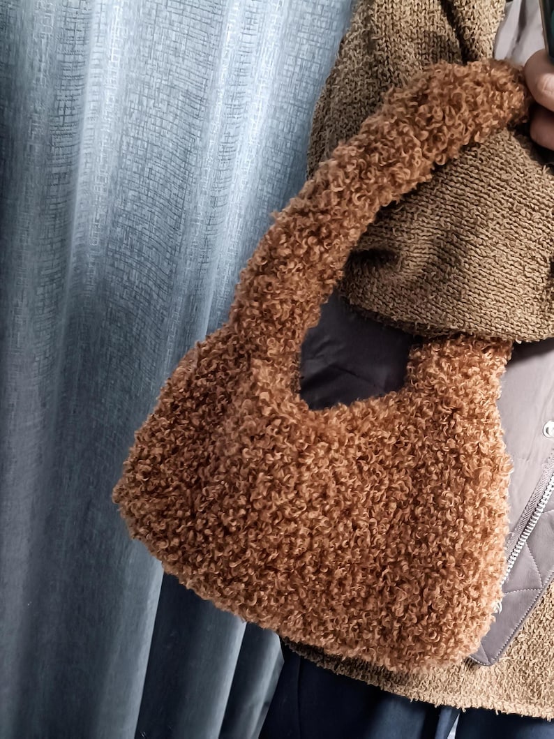 A person holding a brown curly teddy fleece handbag against a blue curtain backdrop.