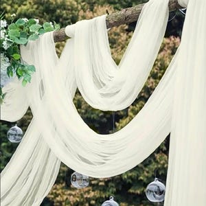 May include: Off-white sheer fabric draped over a wooden branch, creating a flowing, elegant display. Clear glass globes hang below, adding a touch of whimsy. Green foliage and white flowers are visible on the left side, suggesting a wedding or event setting.
