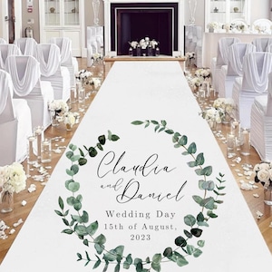 May include: A white wedding aisle runner with the names "Claudia and Daniel" and the date "15th of August, 2023" in a green eucalyptus wreath. The aisle is set up for a wedding ceremony with white chairs and floral arrangements.