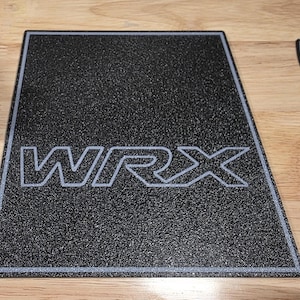 以下が含まれることがあります： ライトグレーの輪郭で「WRX」の文字が入った黒色のテクスチャードマット。 マットにはライトグレーのボーダーと台形があります。 マットは木製の表面にあります。