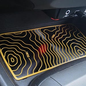 May include: A rectangular black car console mat featuring a gold topographic map design with contour lines and elevation labels reading 1600 and 1800. The mat is positioned in a vehicle storage compartment below a USB port and auxiliary input.
