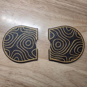 May include: Two black coasters with a gold topographic design. The coasters are semi-circular with a cutout on one side. The design features concentric circles and curved lines, creating a map-like pattern. The coasters are placed on a wooden surface.