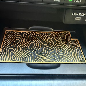 Pode incluir: Um tapete retangular preto para consola de automóvel com um padrão de mapa topográfico dourado de linhas de contorno sinuosas. O tapete está posicionado numa área de armazenamento embutida sob o painel com portas USB visíveis.
