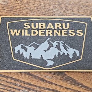 Op de afbeelding: Rechthoekig zwart metalen bord met gouden rand en de woorden "SUBARU WILDERNESS" in gouden letters. Het midden heeft een zilveren bergketen en bomen ontwerp.