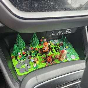 Può includere: Un diorama Lego con una volpe, un ragno e un personaggio di hot dog attorno a un falò. La scena comprende alberi verdi, fiori e pietre grigie su una base verde. Il diorama è posizionato nella console di un'auto.
