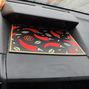 May include: A black rectangular car dashboard mat with a repeating pattern of red chili peppers, green leaves, and small yellow stars. The mat has a gold border and is set in a black dashboard compartment.