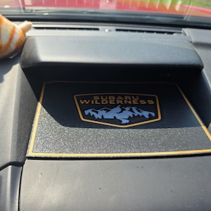 Könnte beinhalten: Schwarzes Armaturenbrett eines Autos mit einem Subaru Wilderness-Logo. Das Logo ist ein goldumrandetes Emblem mit den Worten "Subaru Wilderness" über einer blauen Bergketten-Grafik. Ein kleines orange-weißes Spielzeugtier befindet sich auf dem Armaturenbrett.