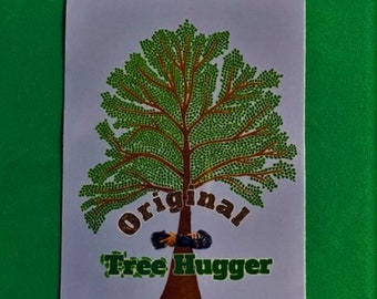 May include: A sticker featuring a stylized tree with a brown trunk and branches, and green leaves. The text "Original Tree Hugger" is printed below the tree in brown and green. The sticker is on a white background, set against a green surface.
