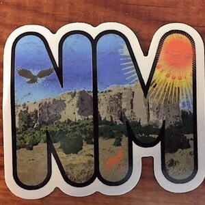 May include: A sticker featuring the letters "NM" with a landscape design. The "N" displays a flying eagle over a mountain range, while the "M" shows a sunburst. The sticker has a white border and a wood grain background.