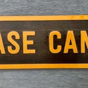May include: A rectangular wooden sign with the words "BASE CAMP" in bold, orange letters. The sign has a dark brown background with an orange border. The sign is likely for a cabin or outdoor setting.