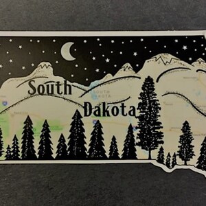 May include: A South Dakota-shaped sticker with a black and white illustration. It depicts a night sky with stars and a crescent moon, a mountain range, and pine trees. The state's name is written across the center.