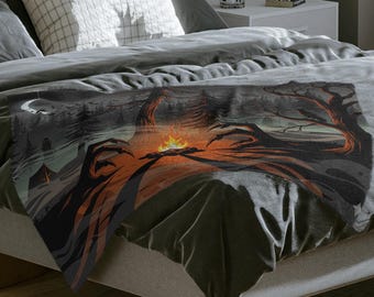 May include: A gray blanket with a dark graphic design featuring a campfire scene with trees and a full moon. The blanket is draped over a bed with gray bedding and pillows. The bed is on a wooden floor.