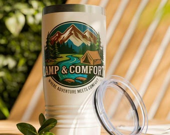 May include: A white insulated tumbler with a camping-themed graphic. The design features a mountain range, river, trees, and a tent, with the words "CAMP & COMFORT" and "WHERE ADVENTURE MEETS COMFORT". A clear lid sits beside the tumbler.