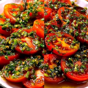 May include: A plate of sliced red tomatoes, each half-covered with a green herb dressing. The tomatoes are arranged on a white plate, and a spoon is placed in the center. The dish is glistening, suggesting it's been drizzled with oil.