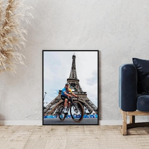 Puede incluir: Una fotografía enmarcada en negro de una persona montando en bicicleta frente a la Torre Eiffel en París, Francia. La persona lleva un maillot de ciclismo azul y amarillo y un casco. La Torre Eiffel está en el fondo y el cielo es azul con nubes blancas.